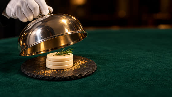 A silver cloche lifts over ivory gaming plaques plated like patisserie on green baize